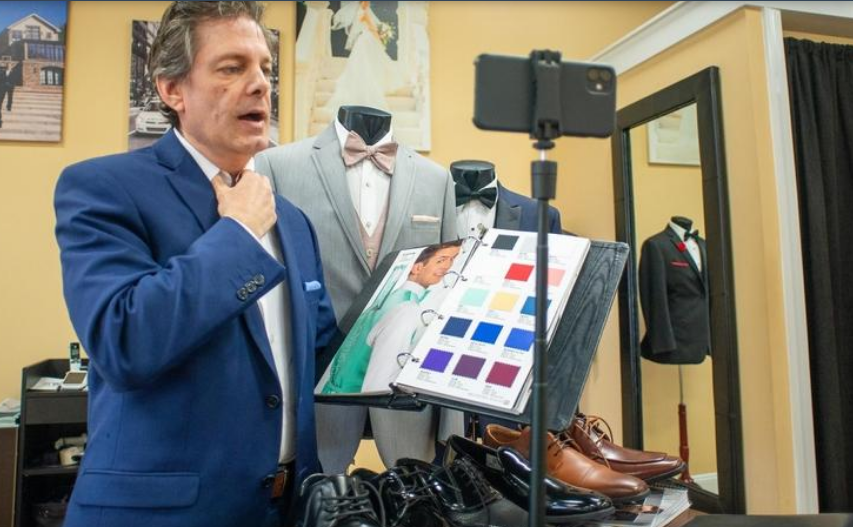 Co-owner of Darianna Bridal & Tuxedo Franco Salerno holds a swatch color book to a video tuxedo appointment, while also displaying  black and brown shoe selections and a gray Michael Kors tuxedo called "Chrome".