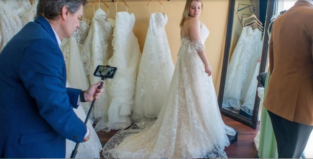 Darianna Bridal & Tuxedo manager Daria Capaldi video models an off-the-shoulder, lace wedding dress with a chapel length train in preparation for an online bridal video appointment