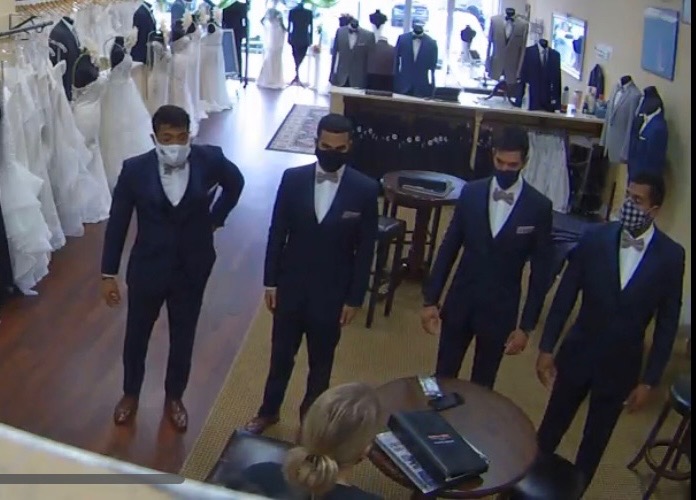 Four groomsmen in Ike Behar navy tuxedos with rose colored bowties are inspected by a Darianna Bridal & Tuxedo staff member. She is making sure they all fit perfectly before they attend the event. A benefit when you rent with us.