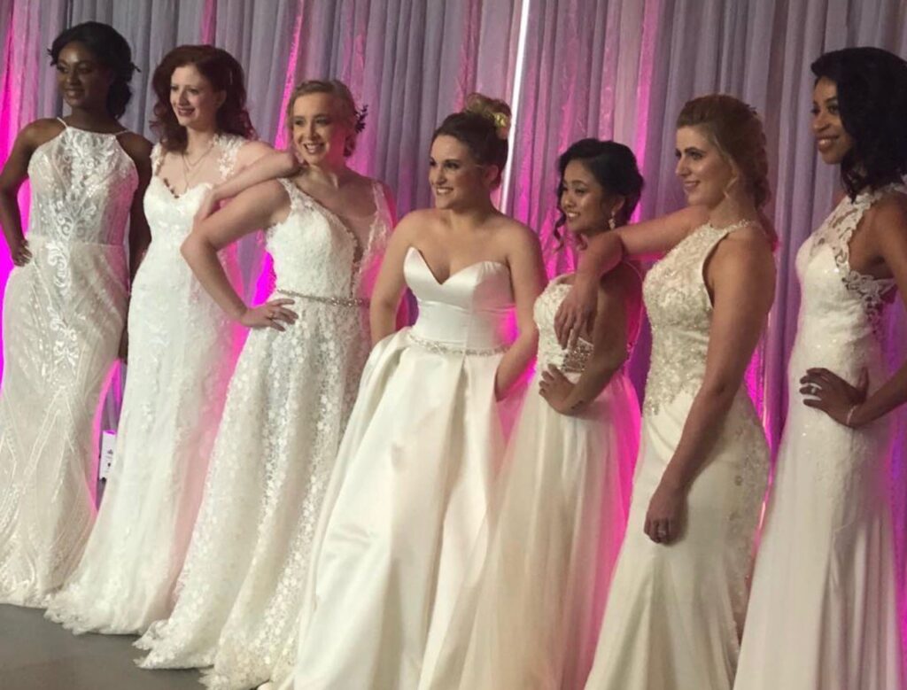 Seven Darianna Bridal & Tuxedo bridal models in various silhouettes of wedding dresses, in fabrics of lace, satin, English net, and crepe, are lined up and ready to walk the runway in a fashion show.