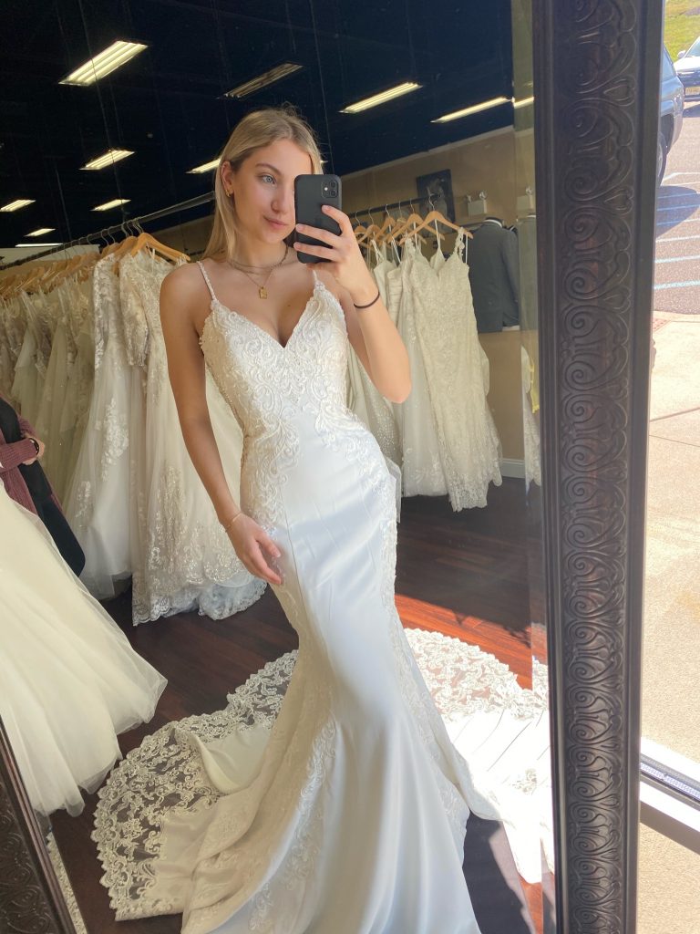 Bride taking a selfie in the mirror wearing a perfectly fitted mermaid dress made of crêpe and lace with a V-neck and beaded straps, and a gorgeous lace train