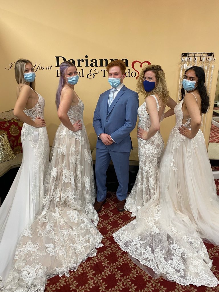 Groom in a cornflower blue color tuxedo and four bridal models looking over their shoulder in lace dresses both fitted and Eileen