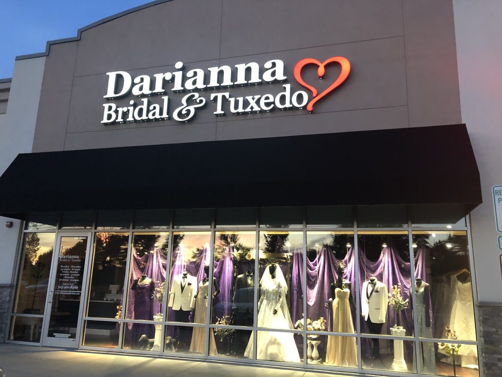 Store front of Darianna Bridal & Tuxedo with wedding dresses and tuxedo mannequins in the window, purple curtains, flowers, white tuxedos lit up at night time. Ready to say yes to the dress!