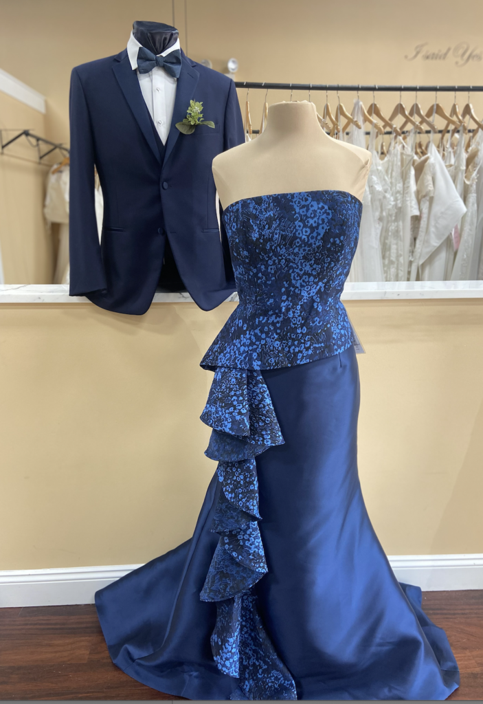 Strapless dress on mannequin with straight across neckline, tight bodice and peplum and front side ruffle. Dark blue and black floral pattern on bodice and ruffle, fit and flare skirt in navy satin.