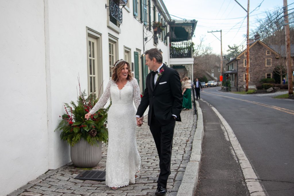 Bride in Madi Lane with Groom in Black tuxedo by Ike Behar walking in front of the black bass hotel in lumberville PA