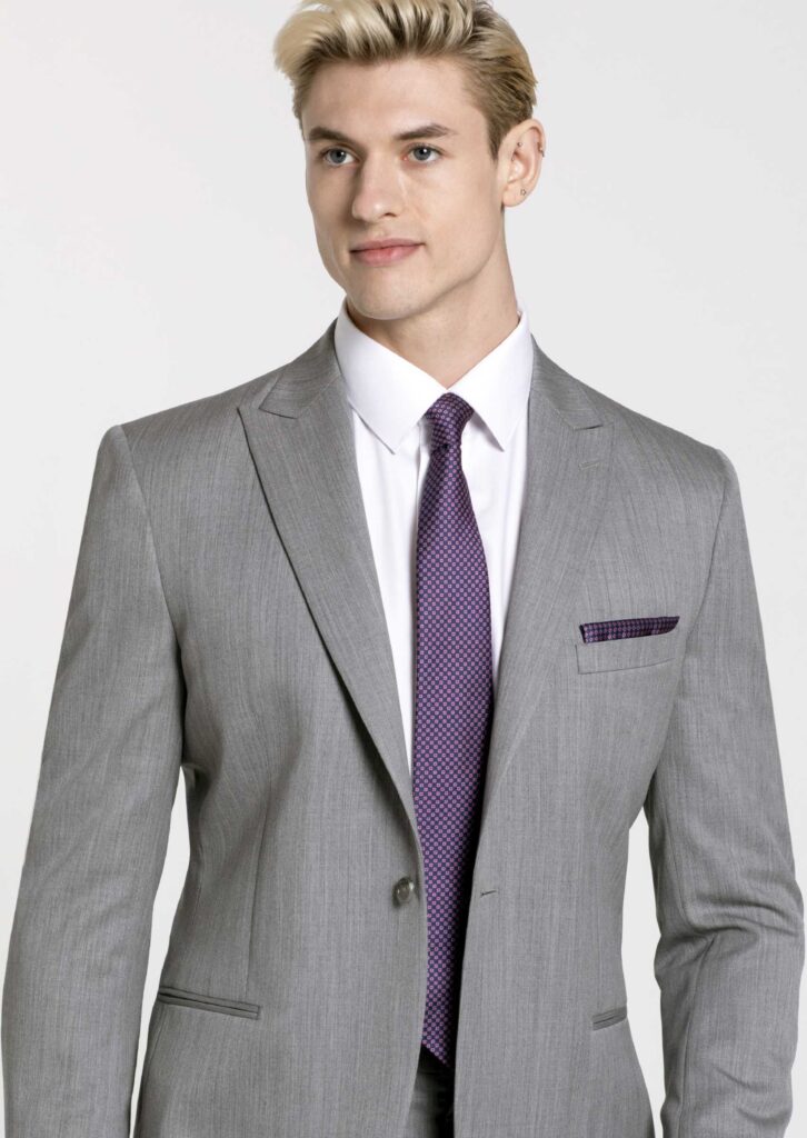Gray suit on model. Heather Gray jacket with peak lapel and one button close. Plum color, long tie and pocket square with checked pattern. White button down shirt.