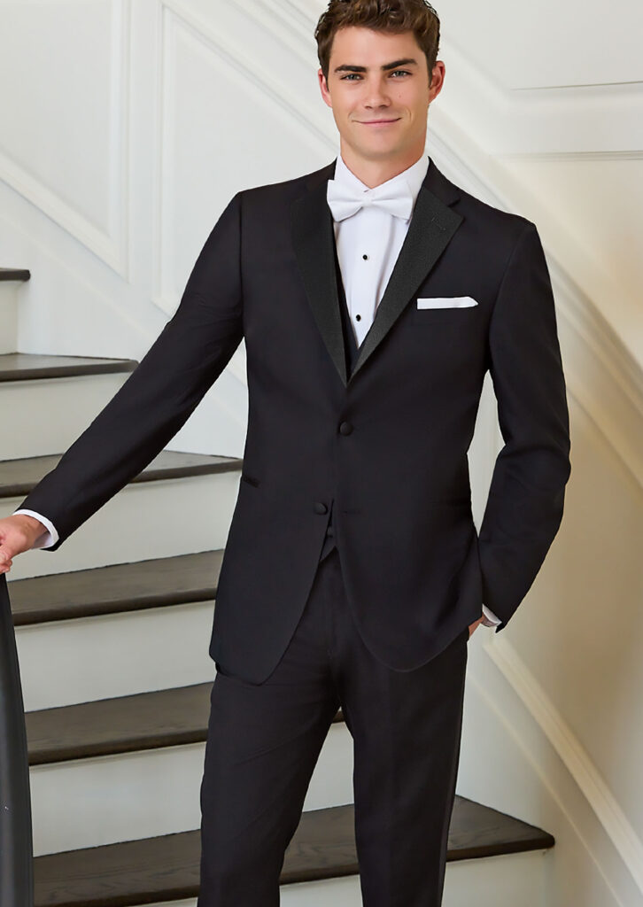 Model in three piece black tuxedo. Satin lapel. White shirt, white bowtie, white pocket square, black button covers. Matching vest and flat front pants.