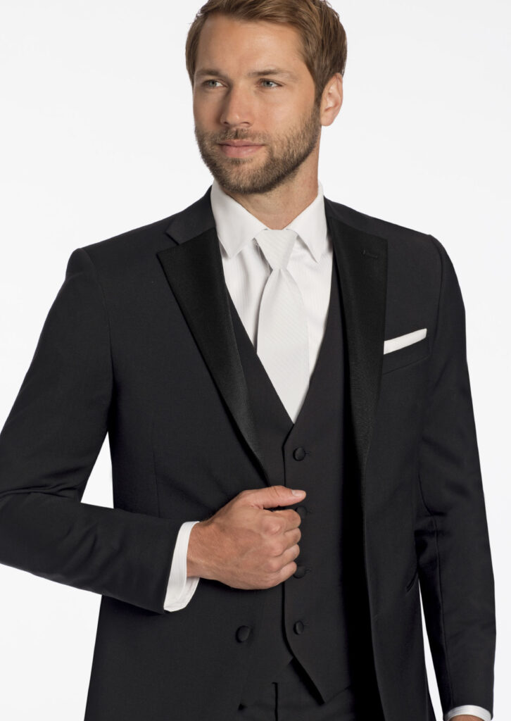 Model in three piece black tuxedo, satin notch lapel jacket, white shirt, white, long tie, white pocket square, matching black vest