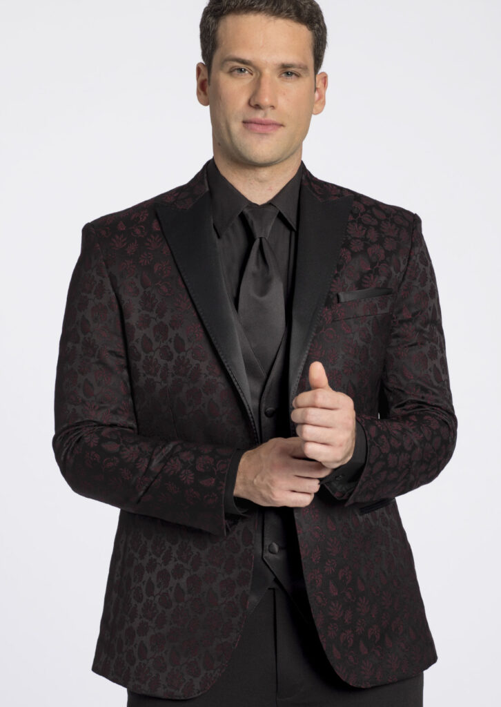 Model in a three piece suit. Black and burgundy, patterned jacket, satin lapel, black shirt, black long tie, black pocket square, black vest, and pants.