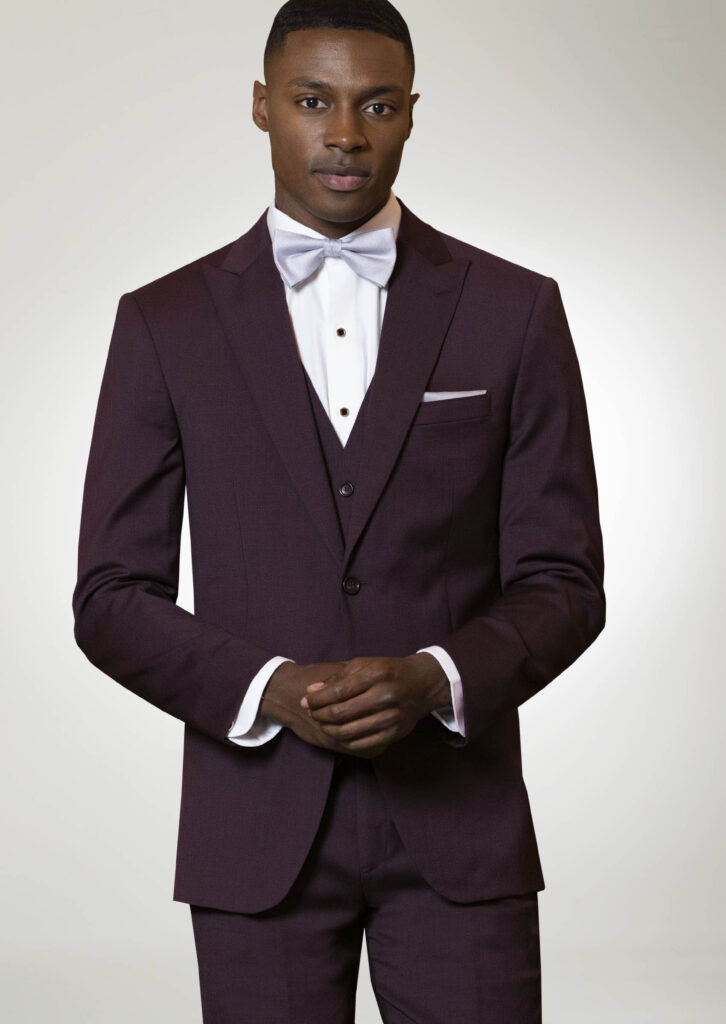 Model in a dark purple suit with a matching vest and pants, lilac bowtie, white shirt with black button covers, lilac pocket square