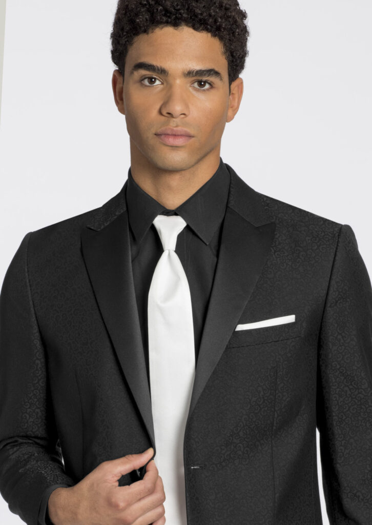 Model in black satin patterned tuxedo jacket, black dress shirt, white satin long tie, white satin pocket square