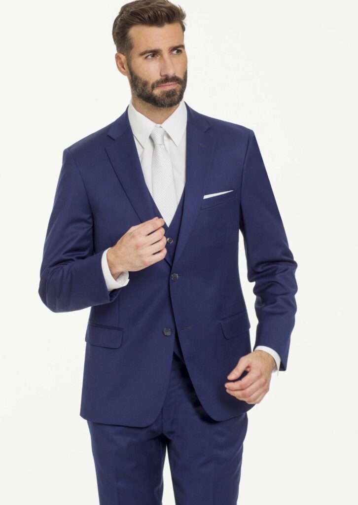 Model in three piece blue suit with white shirt, white long tie, white pocket square, matching blue flat front pants