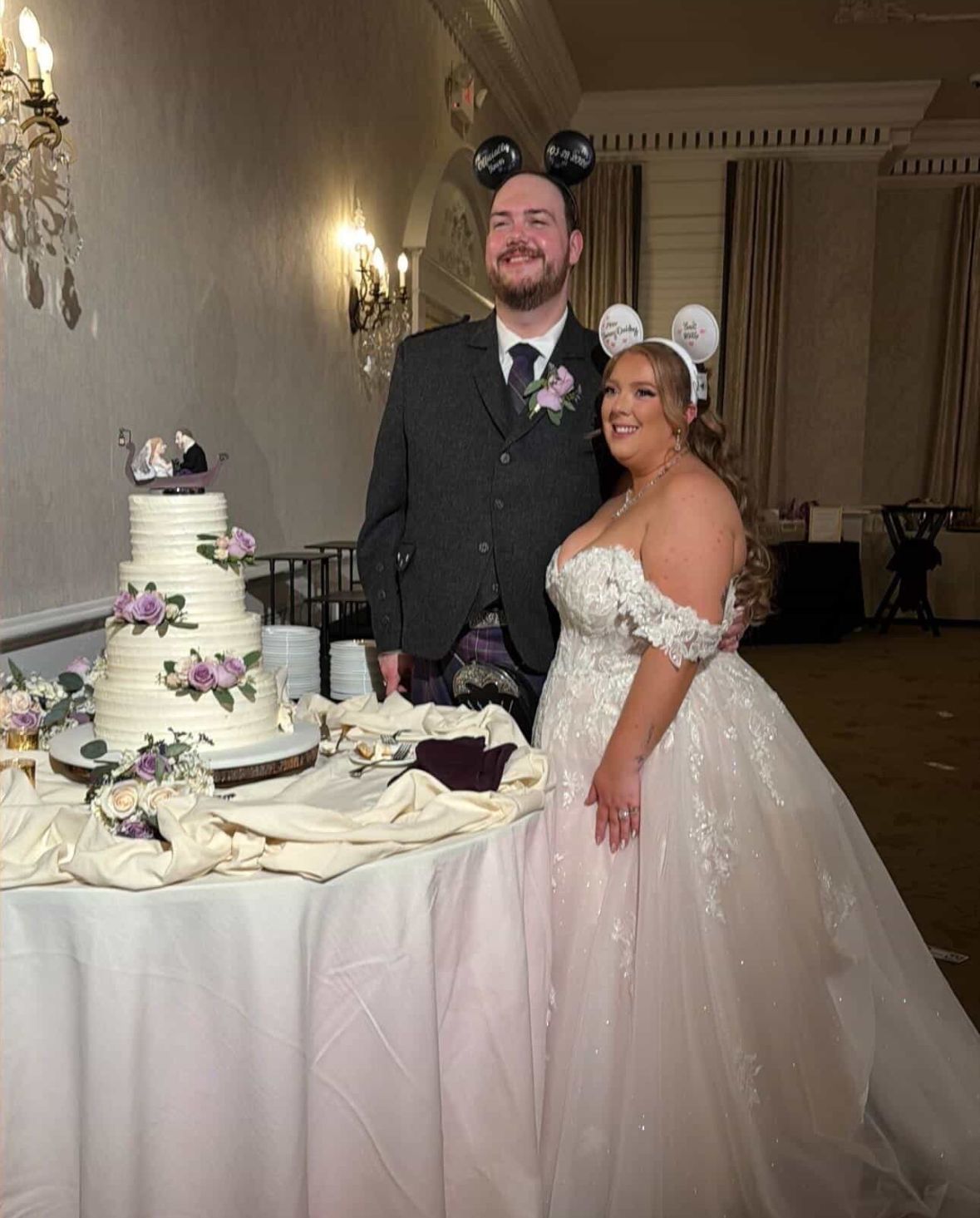Bride and groom standing beside their wedding cake, with the bride wearing an altered off-the-shoulder lace wedding gown